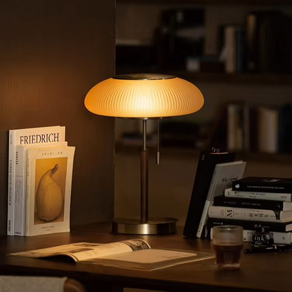 A modern table lamp with a ribbed, mushroom-shaped shade glowing with warm light, set on a dark wooden desk next to books and an open notebook in a cozy room setting.