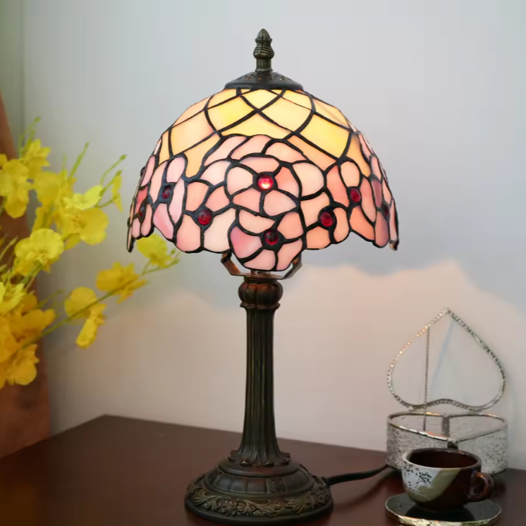 A Tiffany-style stained glass table lamp with a floral pattern in pink, yellow, and red, mounted on an ornate dark metal base and set on a wooden table next to a cup and flowers.