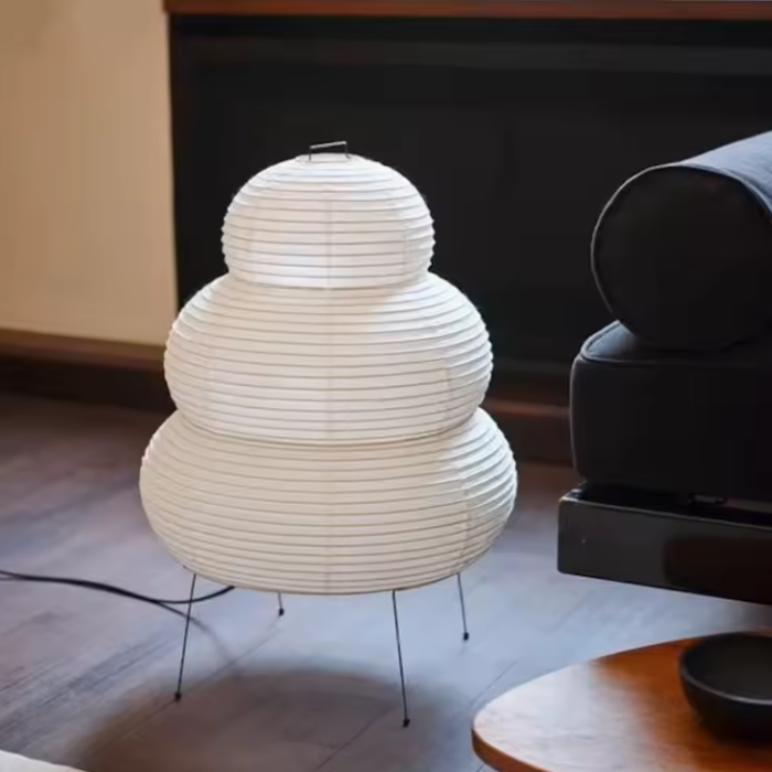 A three-tiered Wabi Sabi Organic Stacked Paper Desk Lamp celebrating Imperfect Natural Textures white paper lantern table lamp with thin metal legs, placed on a dark wood floor in a modern living room setting.