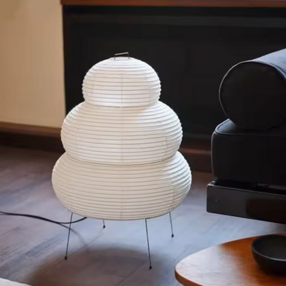A three-tiered Wabi Sabi Organic Stacked Paper Desk Lamp celebrating Imperfect Natural Textures white paper lantern table lamp with thin metal legs, placed on a dark wood floor in a modern living room setting.