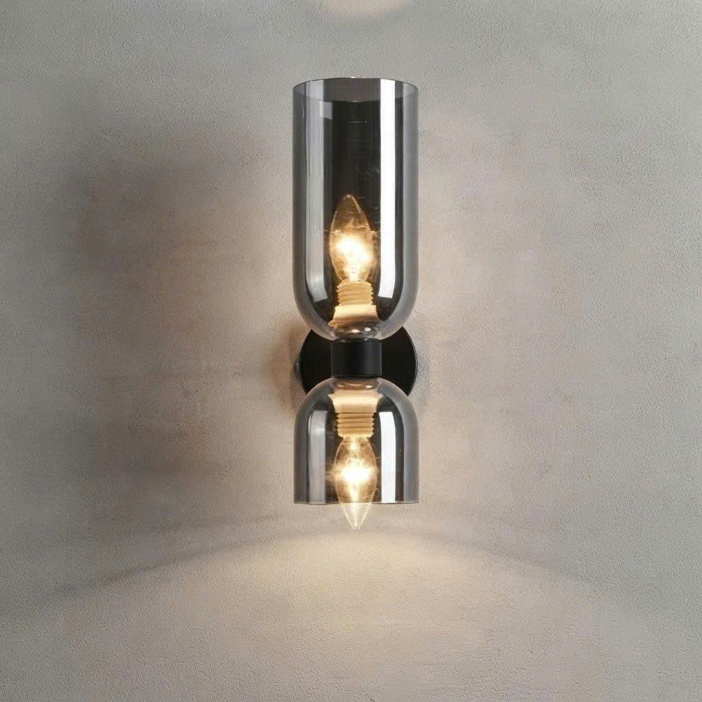 A modern wall-mounted sconce with two mirrored glass shades and glowing warm bulbs, set against a textured light gray wall.