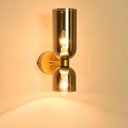 A modern wall-mounted sconce with a brushed brass or bronze metal base and two smoked glass shades, shown lit against a warm beige wall.