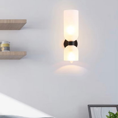 A modern wall-mounted sconce with two frosted glass shades and a black metal center bracket, illuminated against a light gray wall.