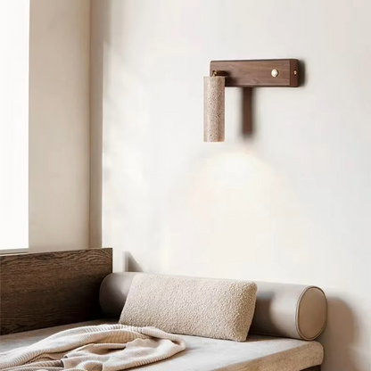 A modern wall-mounted reading light with a dark wood T-shaped bracket and a cylindrical textured lampshade, shown lit above a minimalist beige sofa in a softly styled room.