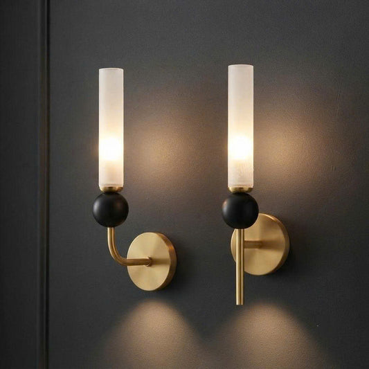 Two modern wall sconces with cylindrical frosted glass shades, matte black spherical accents, and brushed gold circular wall mounts, shown lit against a dark charcoal wall.