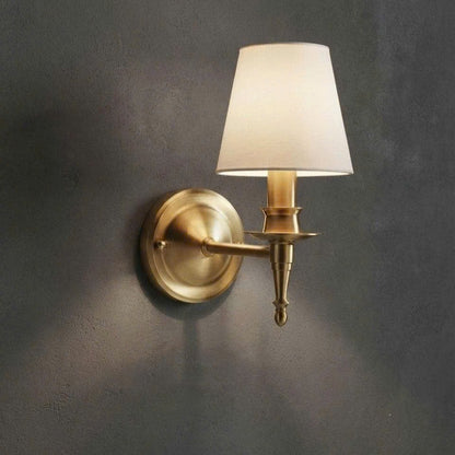 A single wall-mounted sconce with a warm brass/gold metal fixture and a tapered off-white fabric shade, mounted on a dark textured wall.