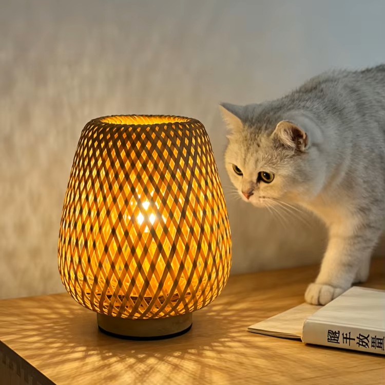 A warm glowing woven table lamp sits on a wooden table next to a gray cat and an open book, with a softly lit neutral wall in the background.