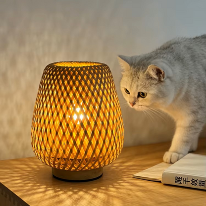A warm glowing woven table lamp sits on a wooden table next to a gray cat and an open book, with a softly lit neutral wall in the background.