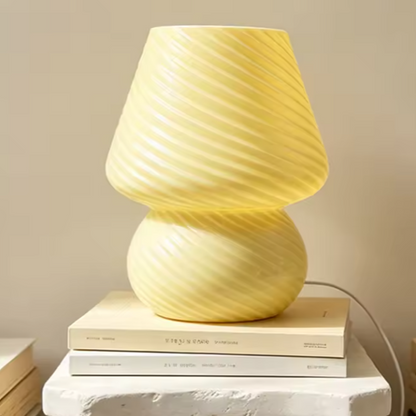 A pale yellow table lamp with a sculptural, ribbed double-bulb design sits on a stack of books on a white stone surface against a warm beige background.