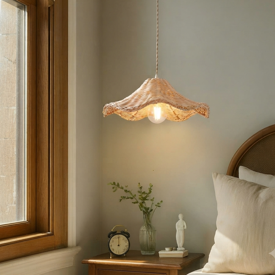 A decorative hand-woven wicker pendant light with a unique wavy petal shape, hanging by a twisted fabric cord above a wooden nightstand in a bedroom setting.