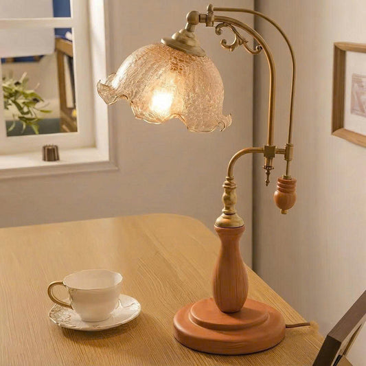 A vintage-style table lamp with a warm glowing textured glass shade, brass-toned metal arms, and a round wooden base, placed on a light wood table in a softly lit room. A white teacup and saucer sit nearby, and a window and framed wall art are visible in the background.