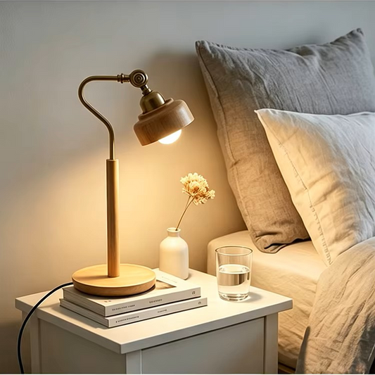 A modern adjustable desk lamp with a light wood base and neck, and a dark metal arm, shown lit on a white bedside table next to a bed with gray and white pillows.