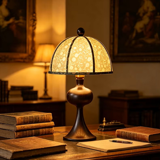 A vintage-style table lamp with a warm glowing stained-glass shade and a dark bronze metal base, placed on a wooden desk in a cozy, dimly lit study with old books and framed artwork in the background.