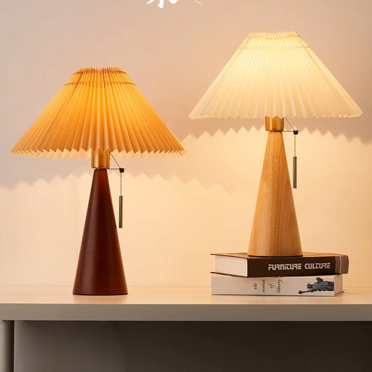Two pleated table lamps on a tabletop against a warm beige wall. The left lamp has a dark reddish-brown tapered base and the right lamp has a light natural wood tapered base, both with cream pleated shades glowing warm yellow when lit.