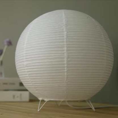 A white spherical paper lantern table lamp with a ribbed texture and three small metal legs, sitting on a light wooden surface against a soft gray background.
